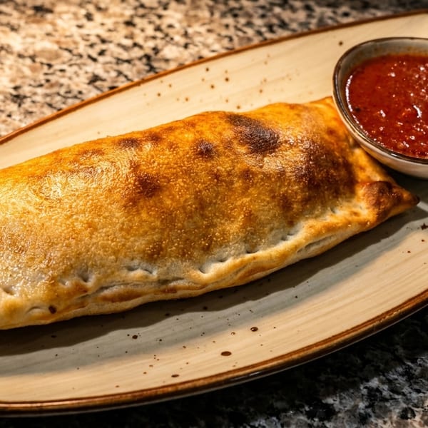 A golden-brown calzone with a side of red marinara sauce on a speckled plate.