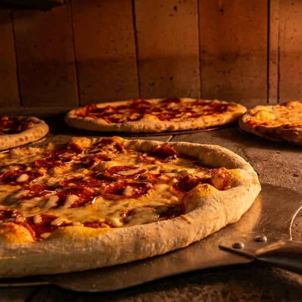 Multiple pizzas, including pepperoni and cheese, baking inside a brick oven with a pizza peel.